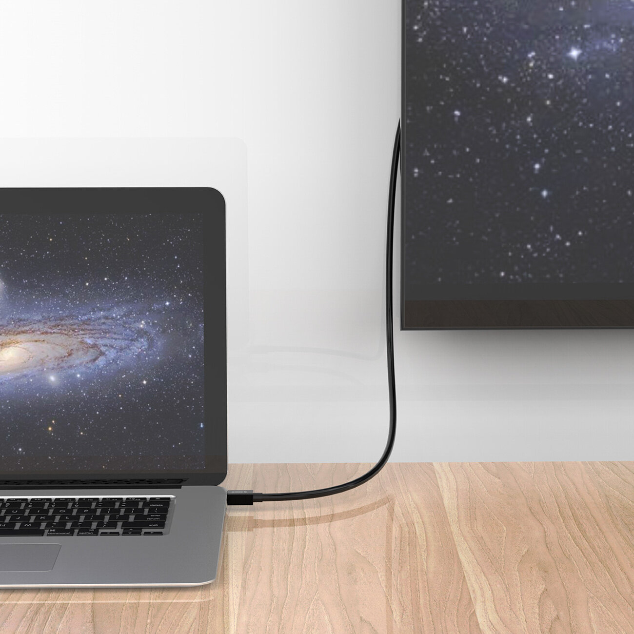 Na drewnianym blacie stoi laptop z wyświetlonym obrazem galaktyki, podłączony kablem do telewizora, na którym widać ten sam obraz.