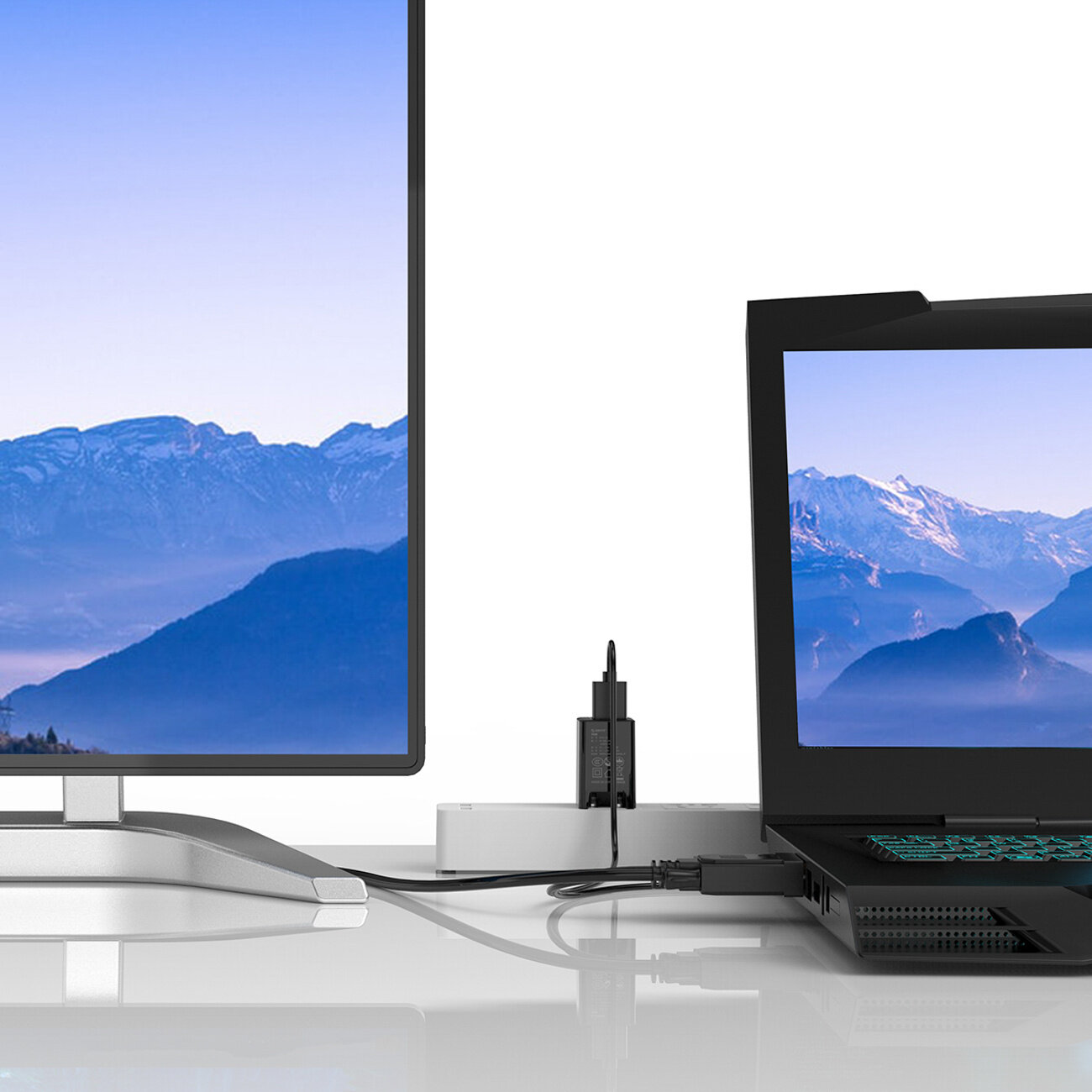 Obraz przedstawia monitor i laptop z wyświetlanym tym samym krajobrazem górskim. Urządzenia są połączone kablem, a w tle widoczny jest adapter z podłączonym zasilaniem USB.