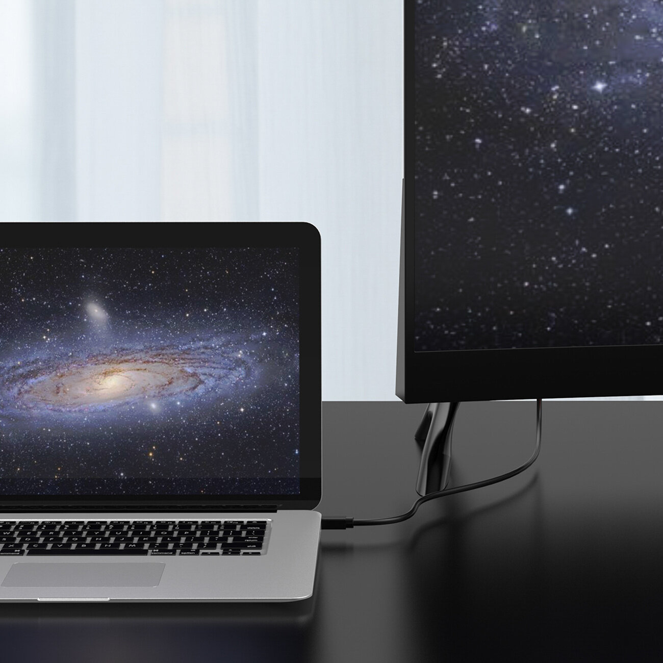 Na biurku znajduje się laptop i monitor, oba wyświetlają obraz galaktyki. Laptop jest podłączony do monitora za pomocą czarnego kabla.