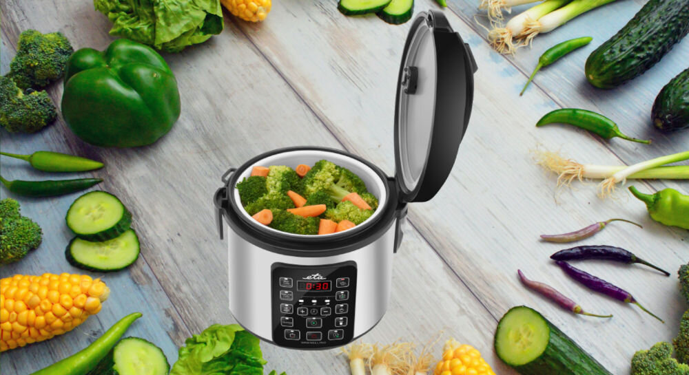 Multicooker z otwartą pokrywą, wypełniony brokułami i marchewką, otoczony różnymi warzywami na drewnianym tle. Na panelu sterowania widoczny jest wyświetlacz z cyframi \'0:30\'.