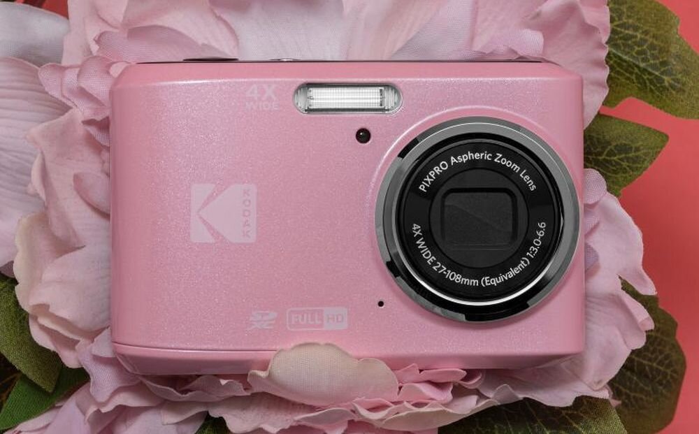 Różowy aparat fotograficzny z logo Kodak, posiadający obiektyw z oznaczeniem \'PIXPRO Aspheric Zoom Lens 4X WIDE 27-108mm (Equivalent) 1:3.6-6.6\'. Aparat leży na tle różowych kwiatów.