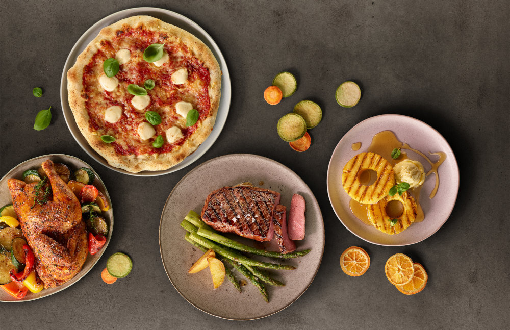 Na obrazie widoczne są cztery talerze z różnymi potrawami: pizza z serem i bazylią, pieczony kurczak z warzywami, stek z szparagami oraz deser z ananasem i lodami. Obok talerzy znajdują się plasterki warzyw i połówki mandarynek.