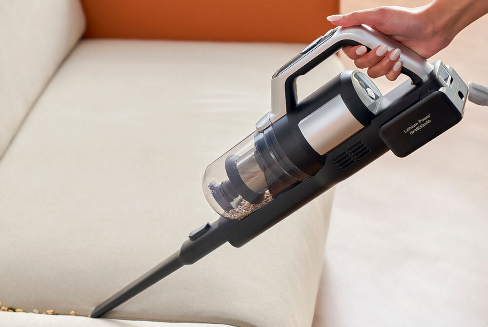 Osoba trzyma odkurzacz ręczny z przezroczystym pojemnikiem na kurz, używany do czyszczenia jasnej kanapy. Na urządzeniu widoczny jest napis \'Lithium Power Cordless Vacuum\'.