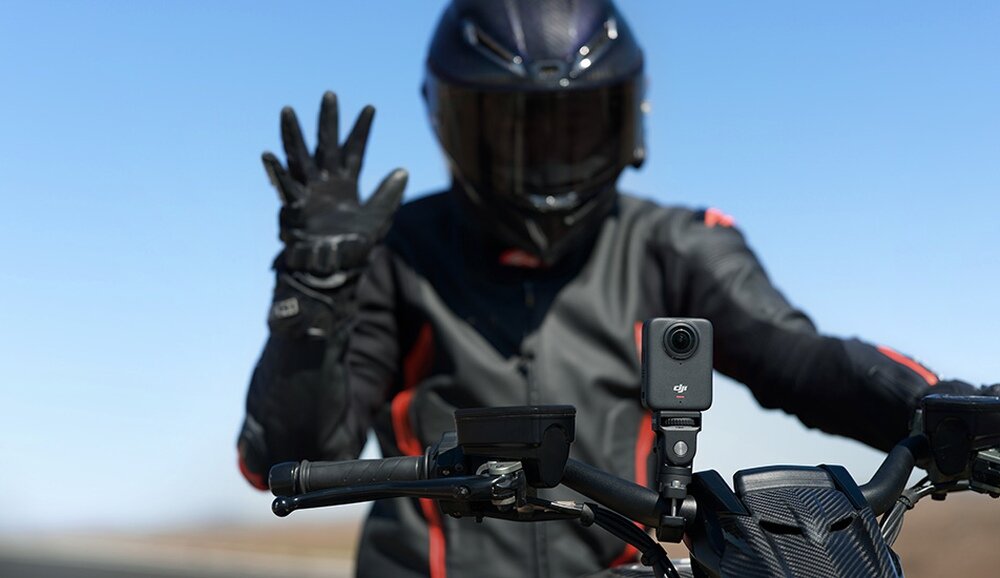 Osoba w kasku motocyklowym i rękawicach trzyma kierownicę motocykla, na której zamontowana jest kamera sportowa z logo DJI.