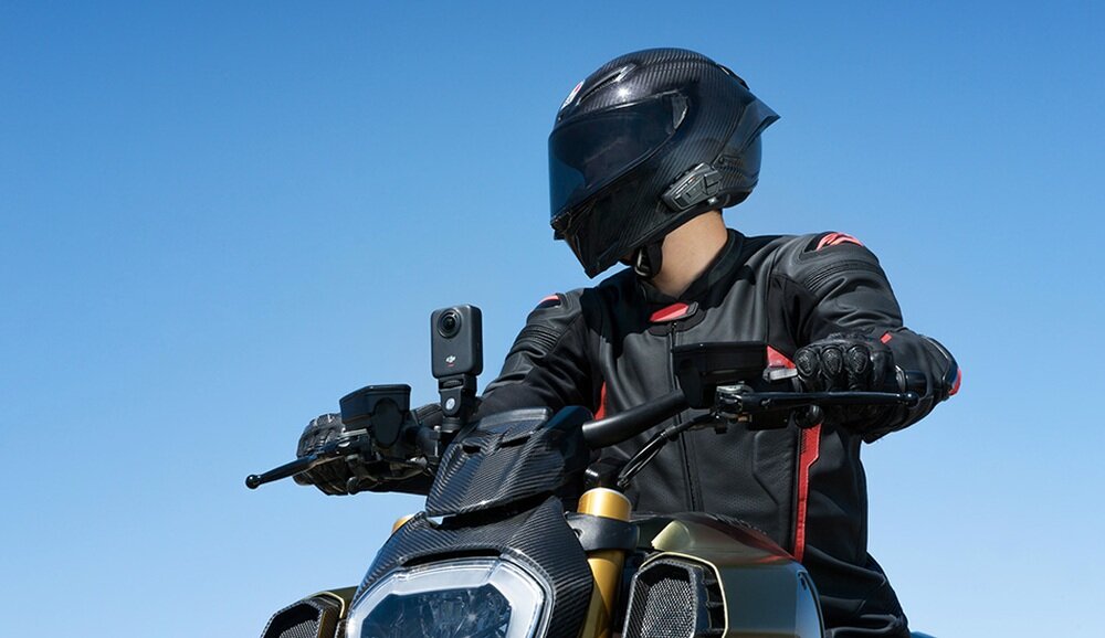 Osoba w kasku motocyklowym i czarnej kurtce siedzi na motocyklu, na którym zamontowana jest kamera sportowa. W tle widać czyste, niebieskie niebo.