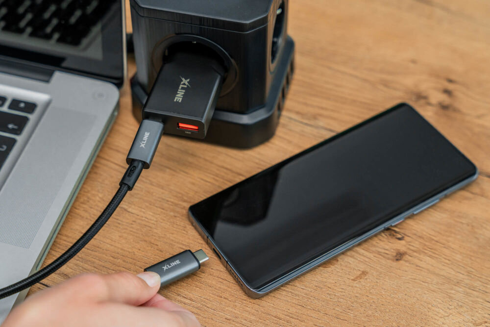 Na drewnianym blacie znajduje się czarny smartfon, laptop oraz ładowarka z logo \'XLINE\'. Osoba trzyma kabel USB-C z logo \'XLINE\', który jest podłączony do ładowarki.