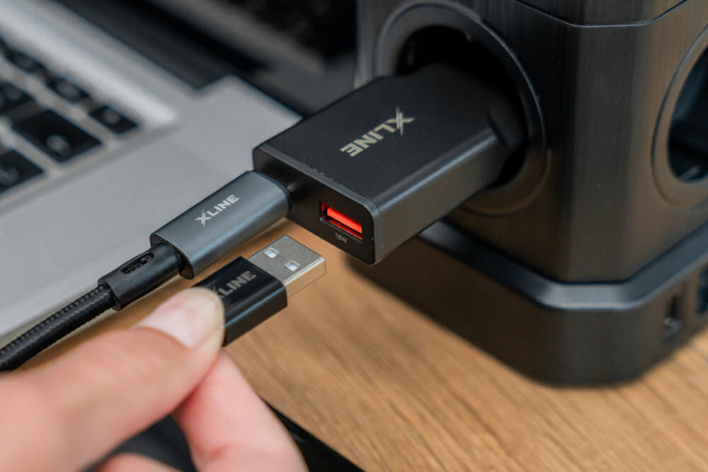 Na obrazie widać czarną ładowarkę sieciową z logo \'XLINE\' podłączoną do gniazdka. Obok znajduje się kabel USB z tym samym logo, trzymany przez osobę.