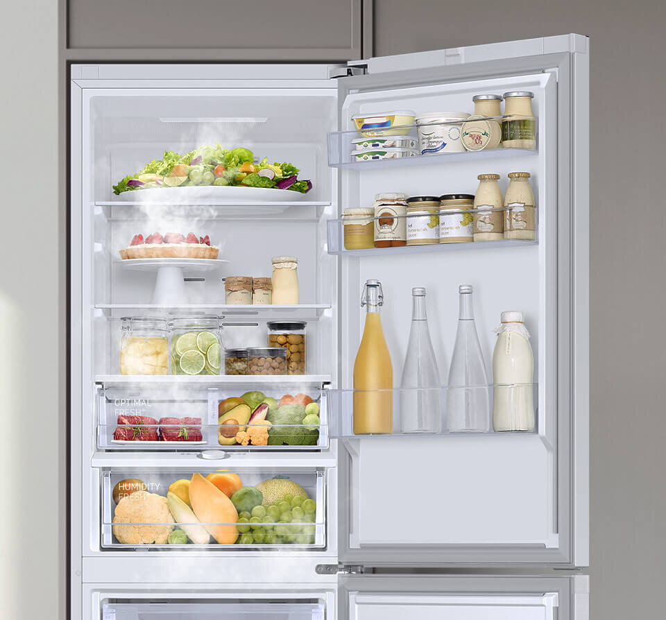 Otwarta lodówka z widocznymi półkami wypełnionymi różnorodnymi produktami spożywczymi, takimi jak owoce, warzywa, napoje i słoiki. Na drzwiach lodówki znajdują się dodatkowe półki z butelkami i słoikami.