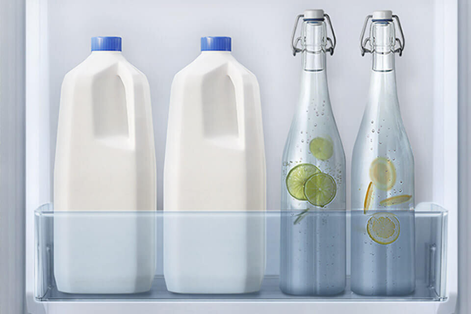 W lodówce znajdują się dwie plastikowe butelki z mlekiem z niebieskimi zakrętkami oraz dwie szklane butelki z wodą i plasterkami cytryny i limonki.