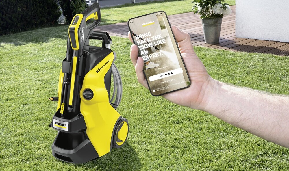 Na trawniku stoi żółto-czarna myjka ciśnieniowa z widocznym logo KARCHER. Obok osoba trzyma smartfon z wyświetlonym tekstem: \'BRING BACK THE WOW LIKE AN EXPERT\'.