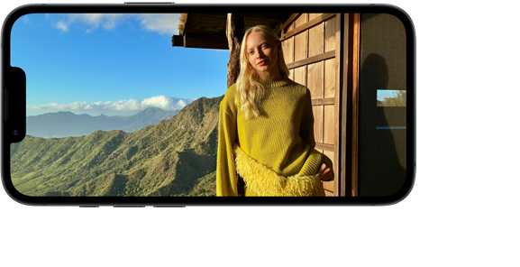 Smartfon wyświetla zdjęcie osoby stojącej na tle górskiego krajobrazu z błękitnym niebem i chmurami. Osoba ubrana jest w żółty sweter, a za nią znajduje się drewniana konstrukcja.
