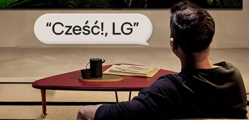 Osoba siedzi przed telewizorem, na ekranie widoczny jest tekst w dymku: \'Cześć!, LG\'. Na stoliku obok znajduje się kubek i książka.