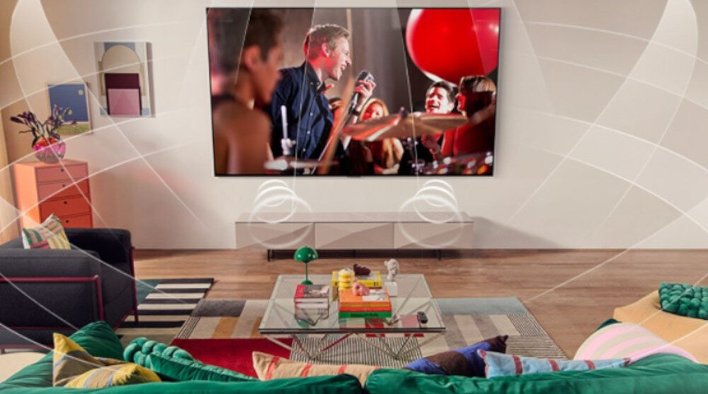 W salonie znajduje się duży telewizor wyświetlający scenę z koncertu. W pomieszczeniu widoczne są kolorowe meble i dekoracje, a na ekranie widać ludzi z mikrofonem i czerwonym balonem.