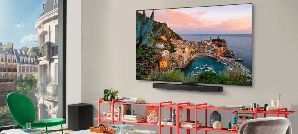 W salonie na ścianie wisi duży telewizor wyświetlający kolorowy krajobraz nadmorskiego miasteczka. Przed telewizorem znajduje się czerwona półka z książkami i dekoracjami, a obok stoi zielone krzesło.