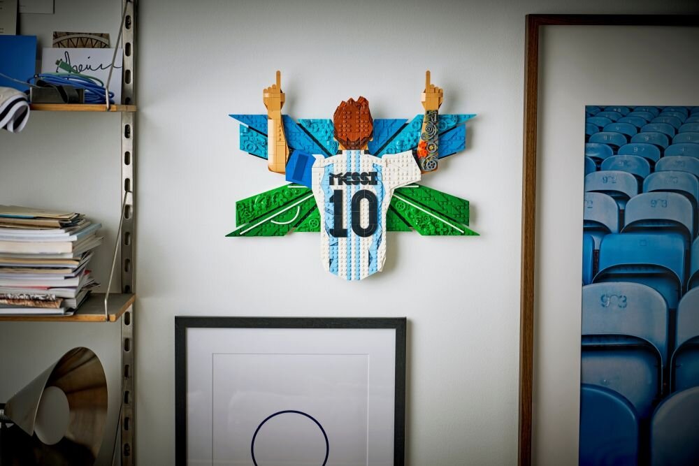 Na ścianie wisi konstrukcja z klocków LEGO przedstawiająca piłkarza z numerem 10 i napisem \'Messi\' na koszulce, z uniesionymi rękami. Obok znajdują się półki z książkami i zdjęcie przedstawiające rzędy niebieskich krzeseł stadionowych.