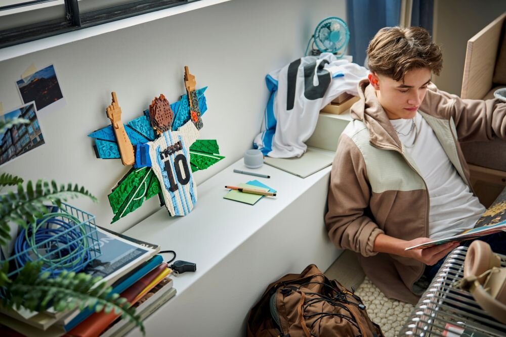Na półce znajduje się model LEGO przedstawiający piłkarza z uniesionymi rękami, obok leży koszulka z numerem 10. Obok siedzi osoba przeglądająca książkę, a na półce są również notatki i kubek.