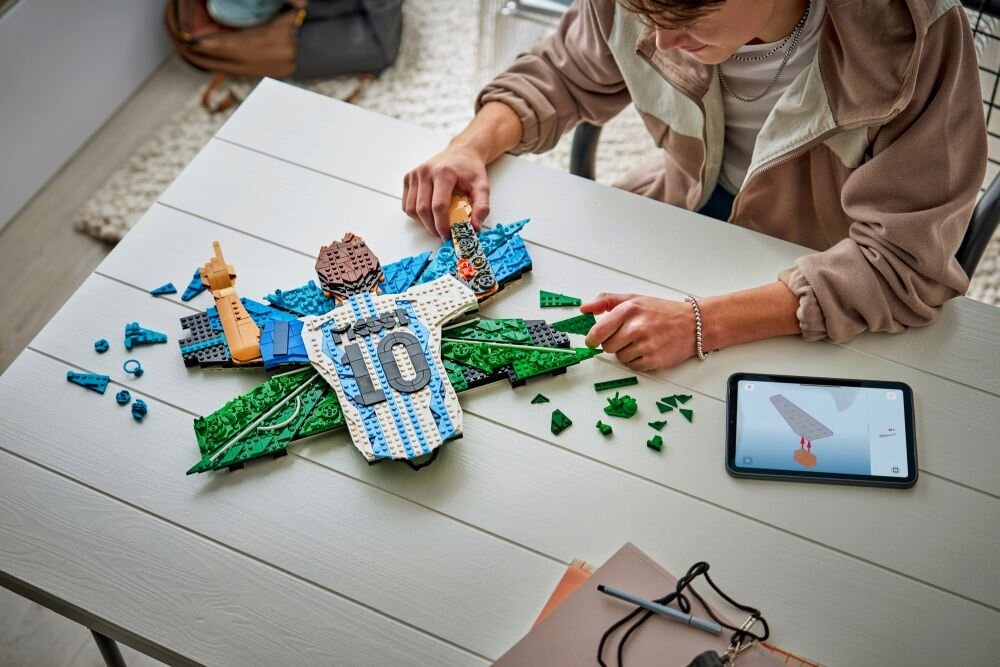 Osoba składa zestaw LEGO przedstawiający piłkarza w koszulce z numerem 10 na stole. Obok leży tablet z instrukcją budowy.
