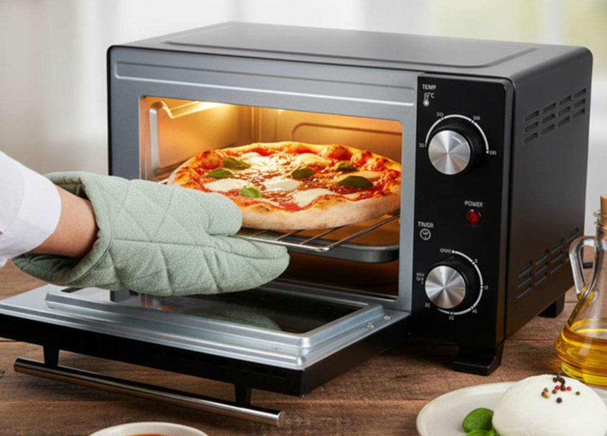 Osoba w rękawicy kuchennej wyciąga pizzę z mini piekarnika. Na panelu piekarnika widoczne są pokrętła z oznaczeniami TEMP i TIMER oraz czerwona dioda POWER.