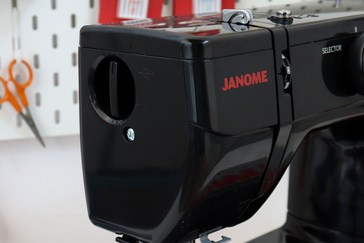 Czarna maszyna do szycia z widocznym logo \'JANOME\' na obudowie. W tle widać nożyczki zawieszone na perforowanej tablicy.