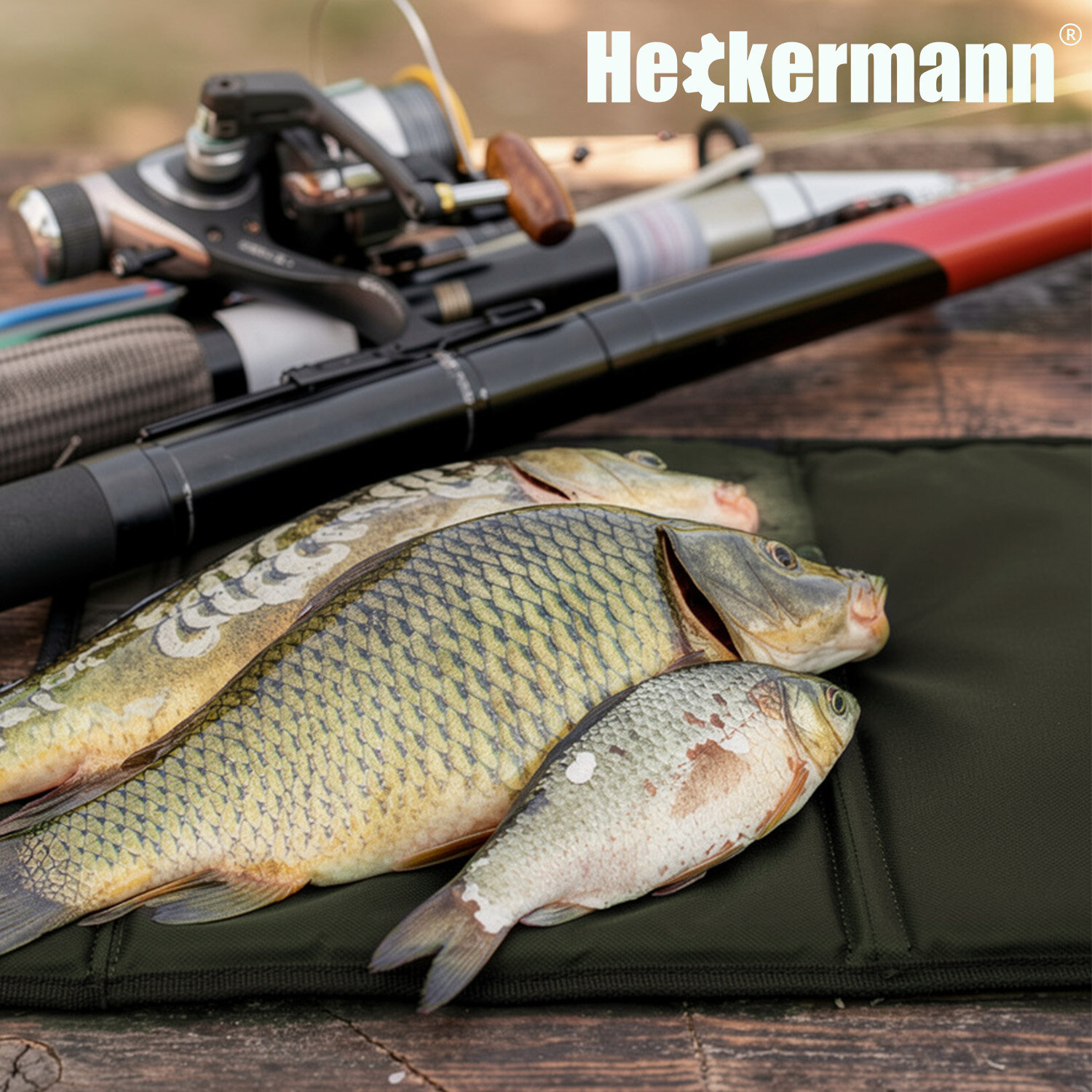 Na ciemnozielonej macie leżą trzy złowione ryby, a obok znajdują się wędki. W górnym rogu widoczne jest logo \'Heckermann\'.
