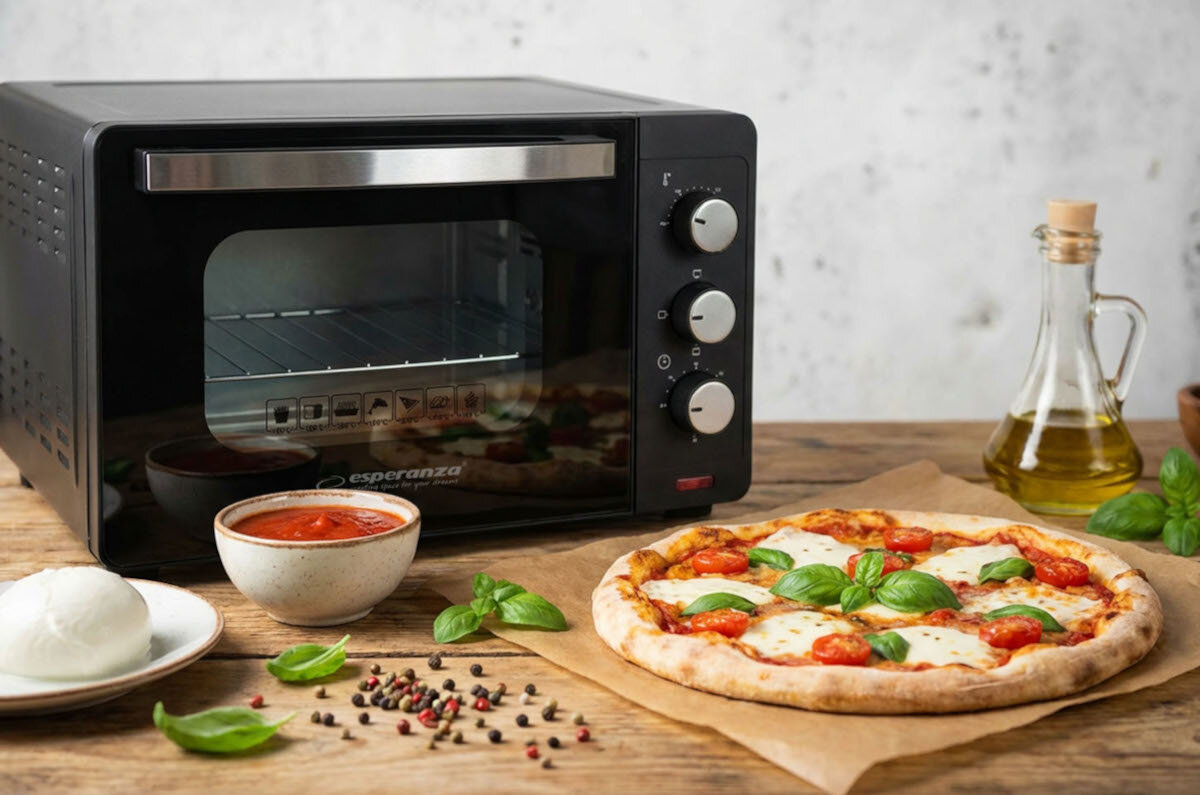 Na drewnianym blacie stoi czarny mini piekarnik z logo \'esperanza\', obok którego znajduje się pizza z mozzarellą i bazylią, miseczka z sosem pomidorowym, kulka mozzarelli, pieprz oraz butelka oliwy.