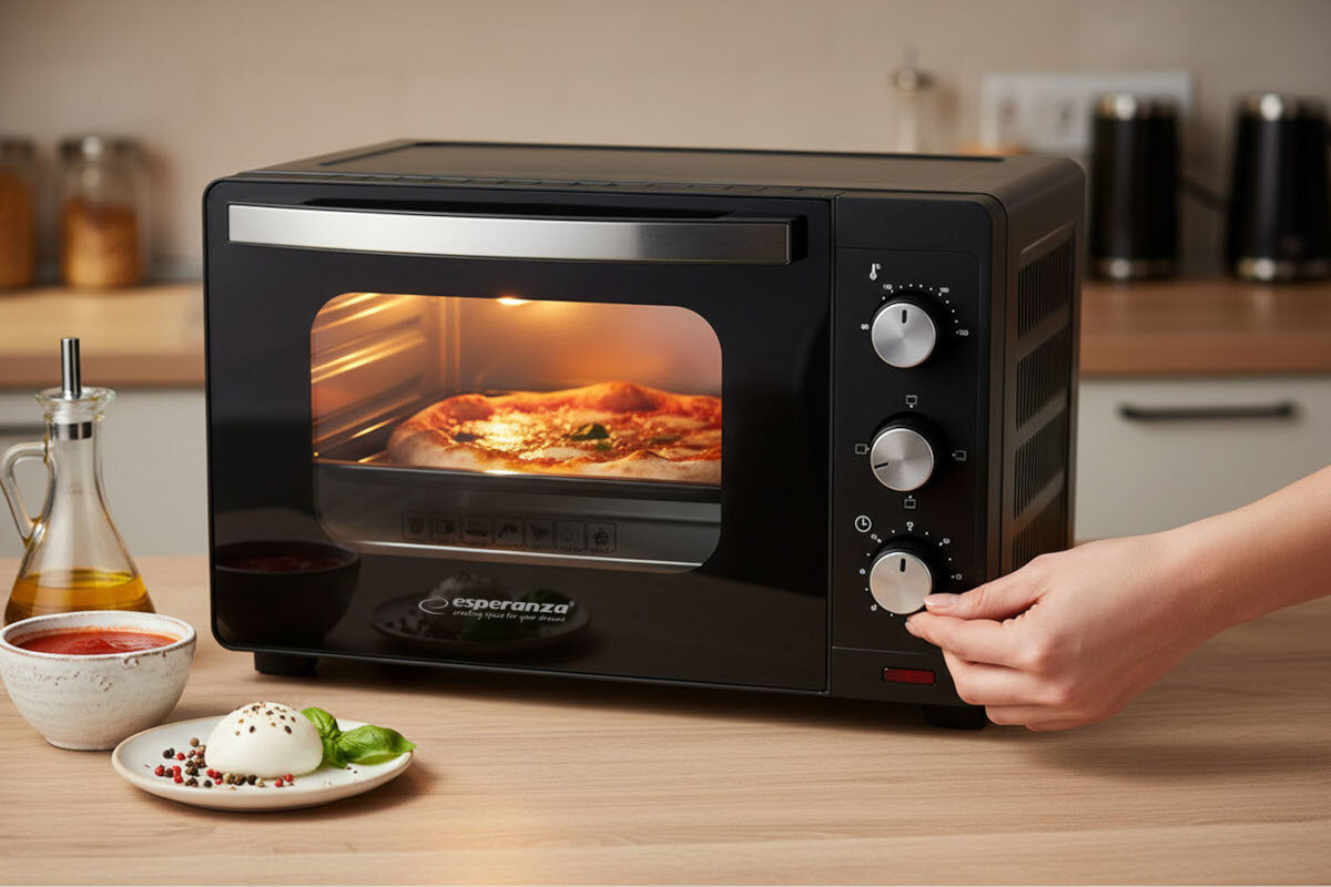 Czarny mini piekarnik z logo \'esperanza\' na froncie, w którym piecze się pizza. Obok piekarnika znajduje się miseczka z sosem, oliwa oraz przyprawy, a ręka reguluje pokrętło na panelu sterowania.