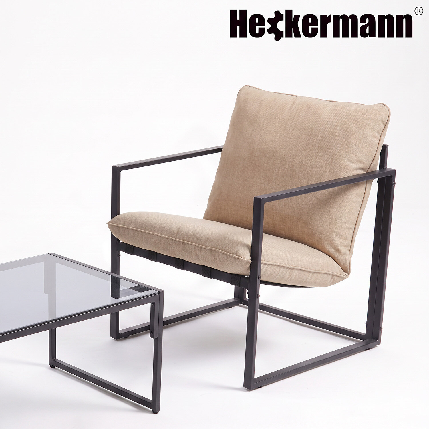 Na obrazie widoczny jest beżowy fotel z metalową ramą oraz szklany stolik. W górnym rogu znajduje się logo \'Heckermann\'.