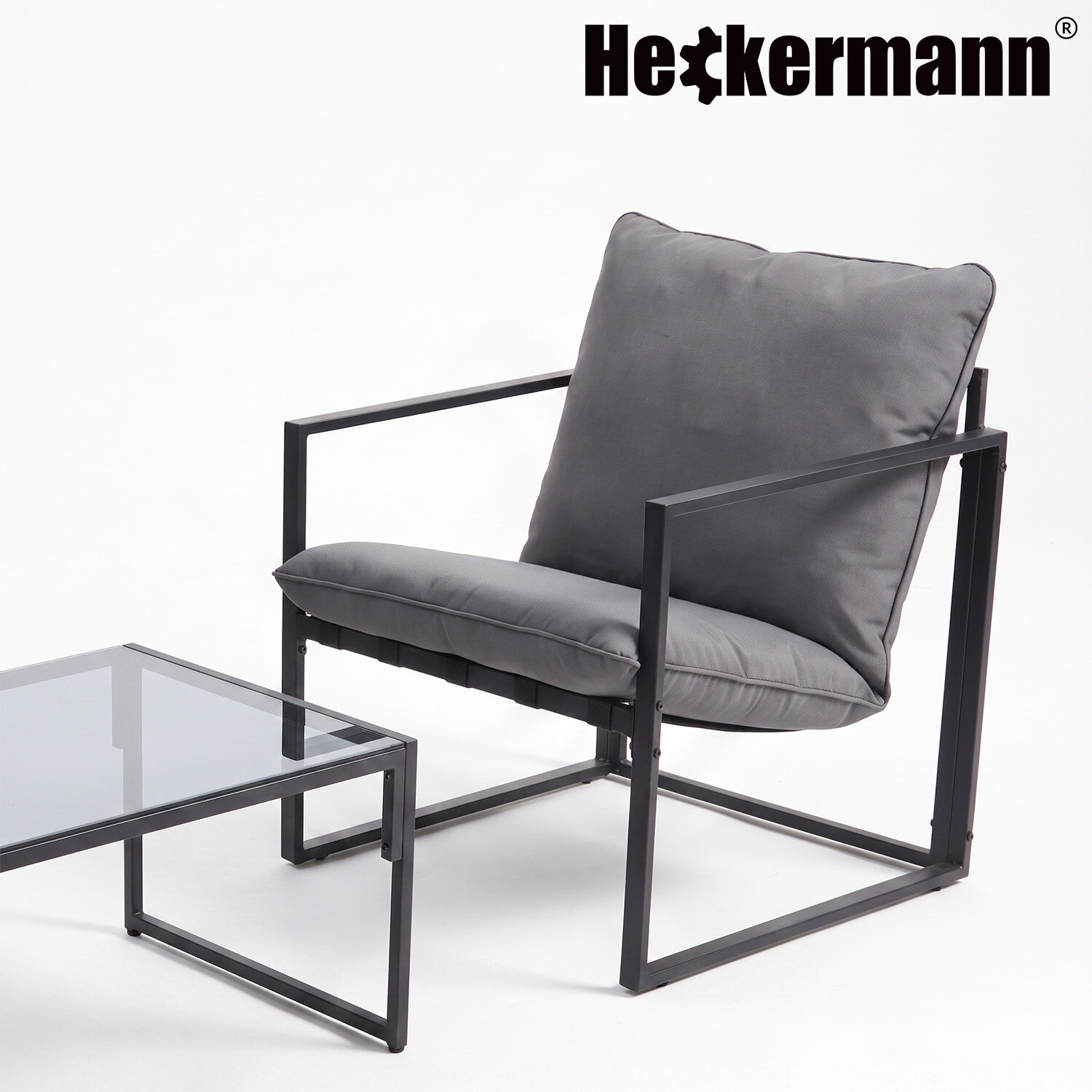 Na obrazie widoczny jest szary fotel z metalową ramą i poduszkami oraz stolik z szklanym blatem. W górnym rogu znajduje się logo \'Heckermann\'.
