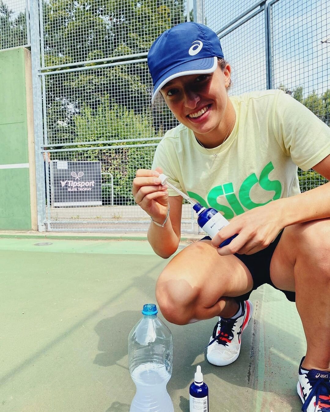 Osoba w czapce i koszulce z logo trzyma butelkę z niebieską etykietą i pipetą. W tle widoczna jest siatka i logo \'Tipsport\'.