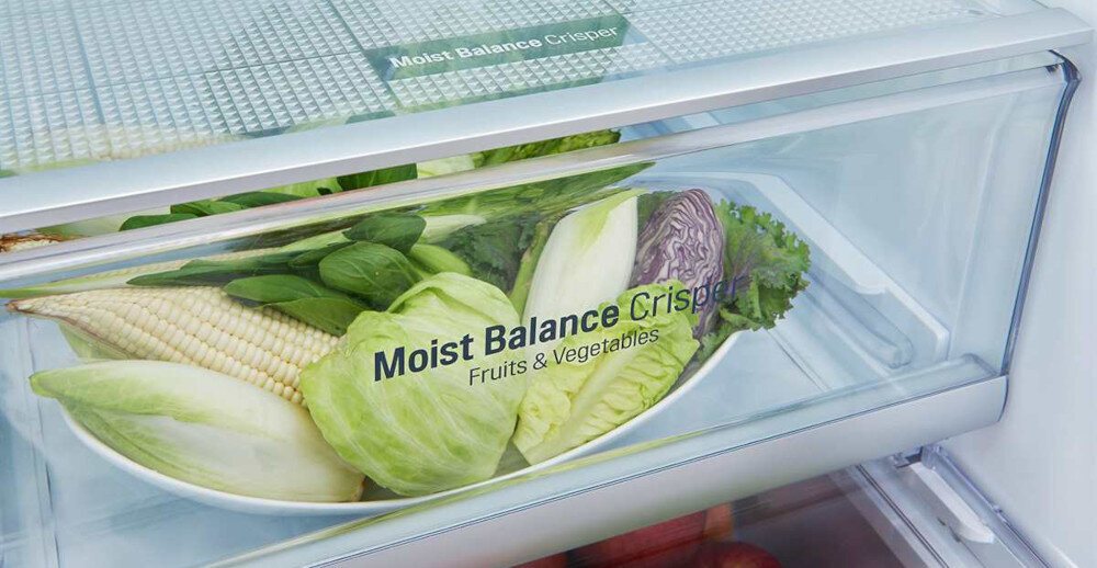 Wnętrze lodówki z szufladą oznaczoną napisem \'Moist Balance Crisper Fruits & Vegetables\', wypełnioną warzywami takimi jak kapusta, kukurydza i sałata. Szuflada ma przezroczystą przednią ściankę.