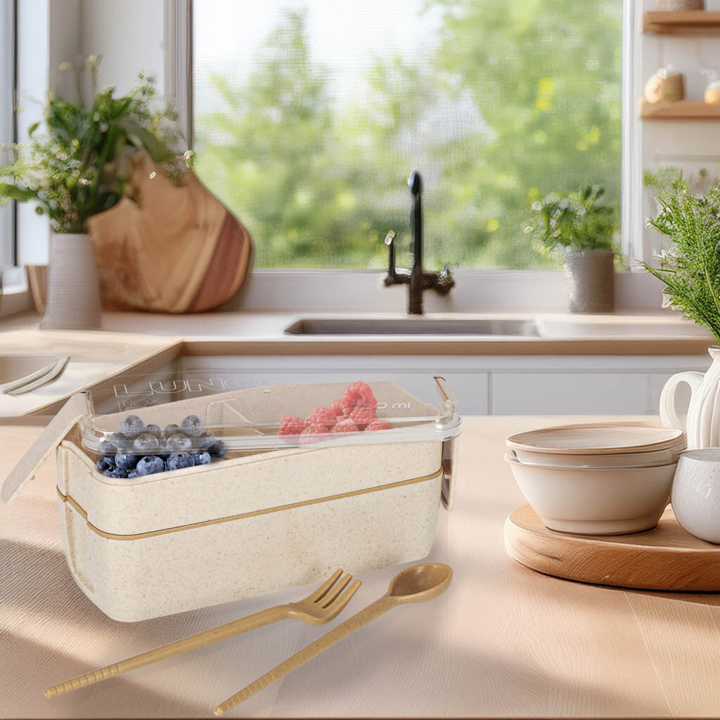 Na blacie kuchennym znajduje się dwupoziomowy lunchbox z przezroczystą pokrywką, wypełniony owocami. Obok leżą złote sztućce, a w tle widać zlew i rośliny doniczkowe.