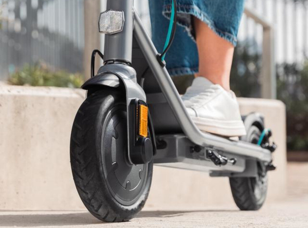Osoba stoi na szarej hulajnodze elektrycznej z dużym przednim kołem i białymi butami sportowymi. W tle widoczny jest fragment betonowej konstrukcji.