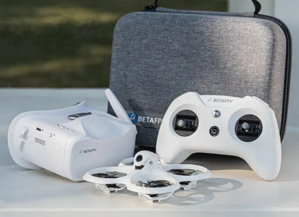 Na zdjęciu znajduje się biały dron z czterema śmigłami, biały kontroler oraz białe gogle, obok szarego etui z logo BETAFPV.