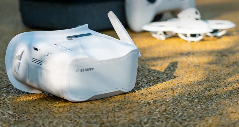 Na ziemi leżą białe gogle z napisem \'BETAFPV\', a w tle widoczny jest biały dron. Oba przedmioty znajdują się na żwirowej powierzchni.