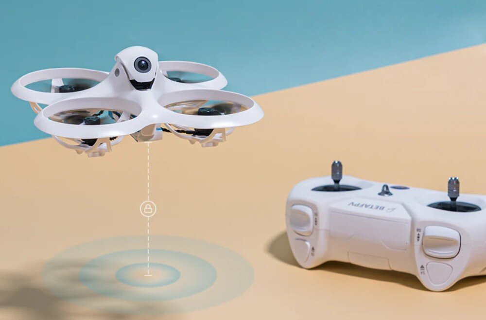 Na obrazie widoczny jest biały dron z czterema śmigłami oraz biały kontroler z napisem \'BETAFPV\'. Dron unosi się nad powierzchnią, a pod nim widoczny jest symbol zasięgu.