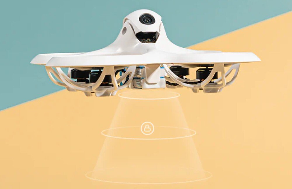 Na obrazie widoczny jest biały dron z kamerą, unoszący się w powietrzu na tle dwukolorowej ściany. Pod dronem znajduje się graficzna reprezentacja sygnału w kształcie stożka z ikoną kłódki.