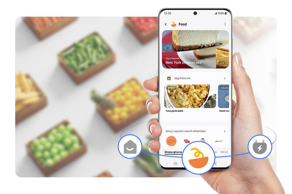 poznaj zakładkę SmartThings Food i polub gotowanie Osoba trzyma smartfon z aplikacją kulinarną, na ekranie widoczny jest przepis na \'New York cheesecake\'. W tle rozmyte pudełka z warzywami.