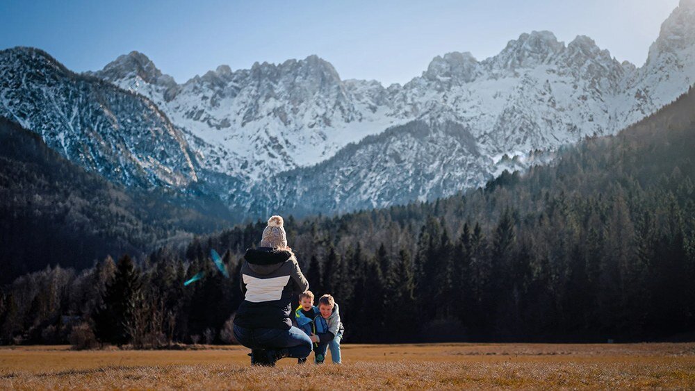 Osoba z dwójką dzieci stoi na polanie z widokiem na ośnieżone góry i las w tle.