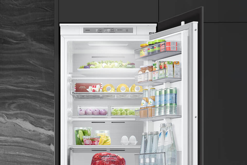 Otwarta lodówka z widocznymi półkami wypełnionymi różnorodnymi produktami spożywczymi, takimi jak warzywa, napoje i jajka. Na drzwiach lodówki znajdują się dodatkowe półki z sokami i puszkami napojów.