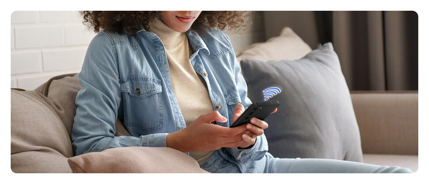 Skorzystaj z aplikacji SmartThings, by zarządzać urządzeniami Samsung z dowolnego miejsca Osoba siedzi na kanapie, trzymając w rękach smartfon z widoczną ikoną sygnału bezprzewodowego. W tle widać zasłony i fragment ściany z cegieł.