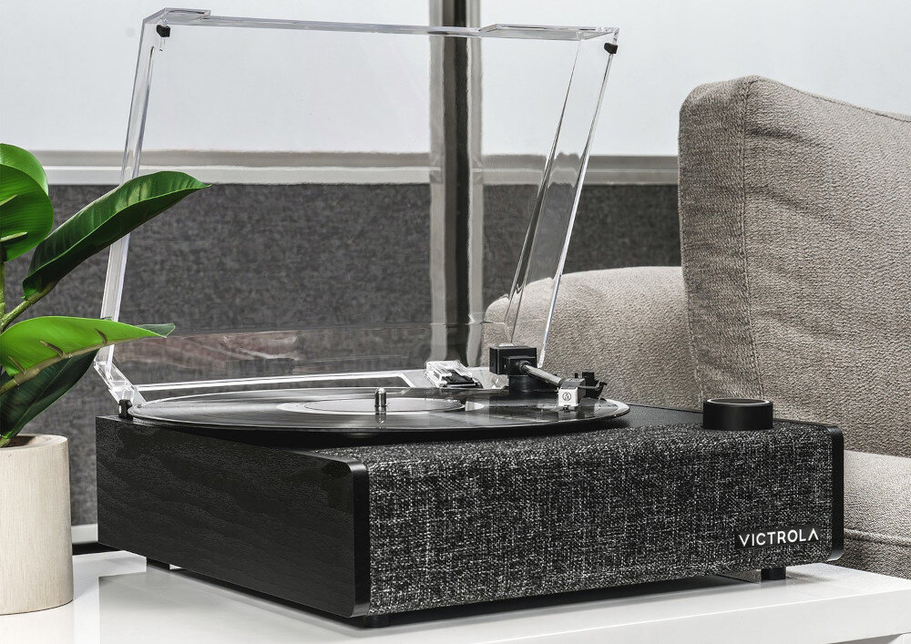 Na obrazie znajduje się czarny gramofon z przezroczystą pokrywą, umieszczony na białym stoliku obok szarej kanapy. Na froncie gramofonu widoczne jest logo \'VICTROLA\'.