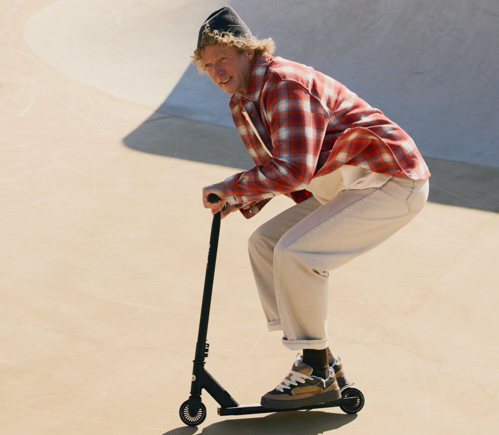 Osoba w czapce i koszuli w kratę jeździ na czarnej hulajnodze wyczynowej po skateparku. Hulajnoga ma czarne koła i prostą kierownicę.