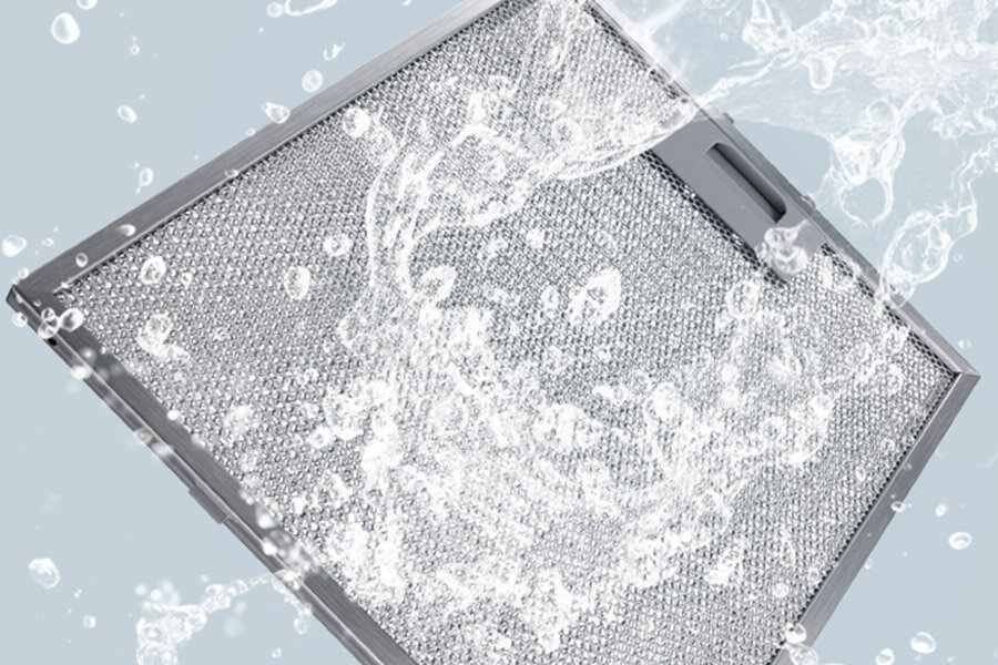 Filtr aluminiowy ułatwia utrzymanie okapu kominowego Samsung NK24C5703TM w czystości Metalowy filtr okapu jest pokazany w trakcie mycia wodą, z widocznymi kroplami i strumieniami wody na jego powierzchni.