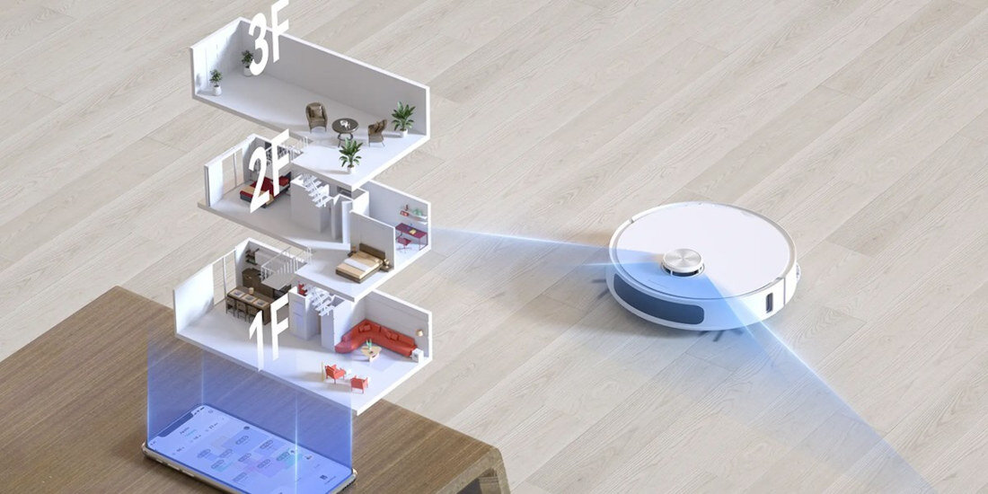 Na drewnianej podłodze znajduje się biały robot sprzątający, emitujący niebieskie światło. Obok widoczny jest model trójwymiarowy trzech pięter domu oraz tablet z aplikacją.