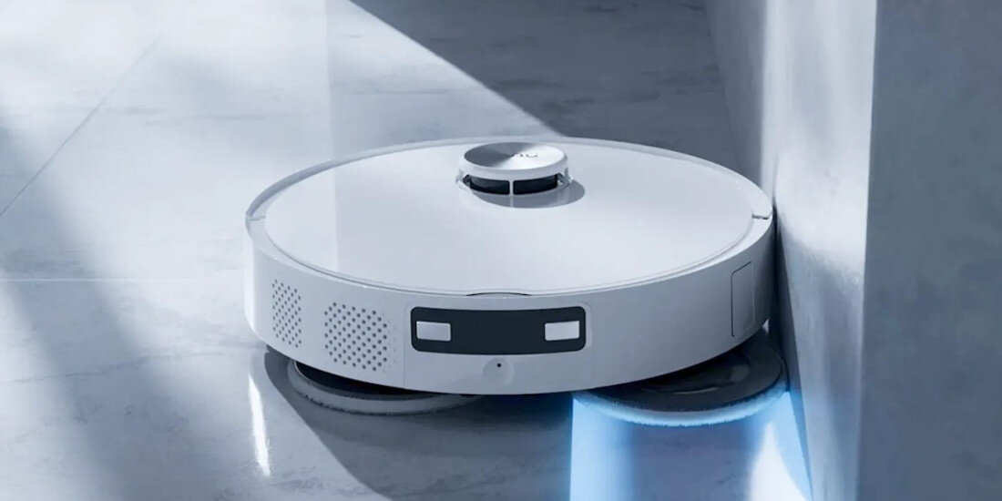 Na obrazie widoczny jest biały robot sprzątający poruszający się wzdłuż ściany. Urządzenie ma okrągły kształt i emituje niebieskie światło od spodu.