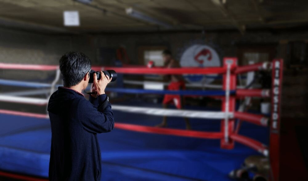 Osoba fotografuje boksera w ringu bokserskim. Na linach ringu widoczne jest logo \'RINGSIDE\'.