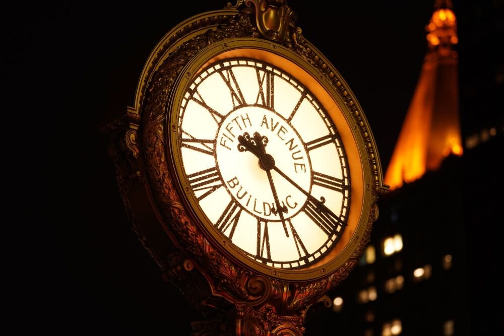 Zegar z rzymskimi cyframi na tarczy, z napisem \'FIFTH AVENUE BUILDING\'. W tle widoczne są oświetlone budynki nocą.