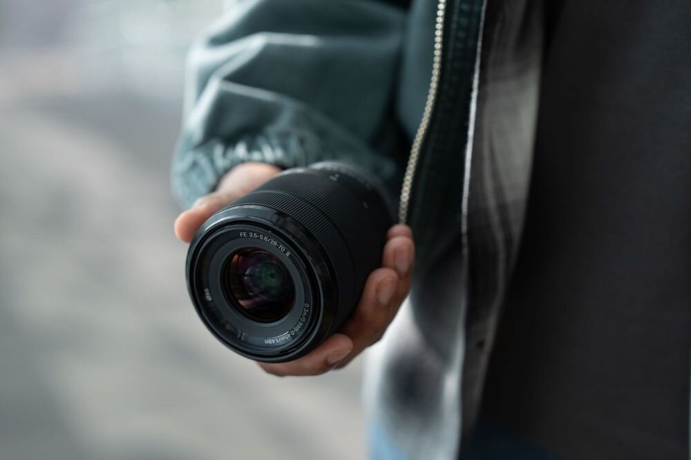 Osoba trzyma obiektyw aparatu fotograficznego w dłoni. Na obiektywie widoczny jest napis \'FE 28-70mm f/3.5-5.6\'.