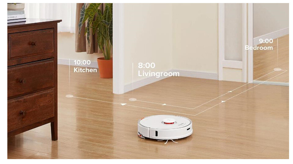 Na drewnianej podłodze znajduje się biały robot sprzątający. Na ścianie widoczne są napisy: \'10:00 Kitchen\', \'8:00 Livingroom\', \'9:00 Bedroom\'.