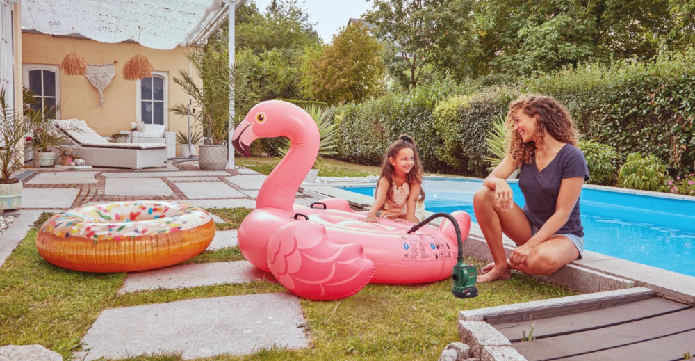 Na trawniku przy basenie znajduje się duży, różowy dmuchany flaming oraz dmuchane koło w kształcie pączka. Obok siedzi osoba z pompka akumulatorową BOSCH EasyInflate 18V-500.